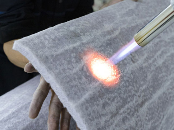 A person holds a thick, white insulating fabric while a blowtorch flame is applied to its surface. The fabric glows red-hot at the contact point, but the person's hands remain unaffected, demonstrating the material’s heat resistance.