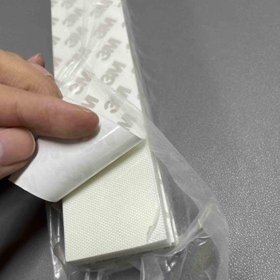 A person peels back the adhesive backing of a white, textured insulation strip with 3M branding, revealing its sticky surface. The strip is partially wrapped in clear plastic packaging and placed on a grey surface.